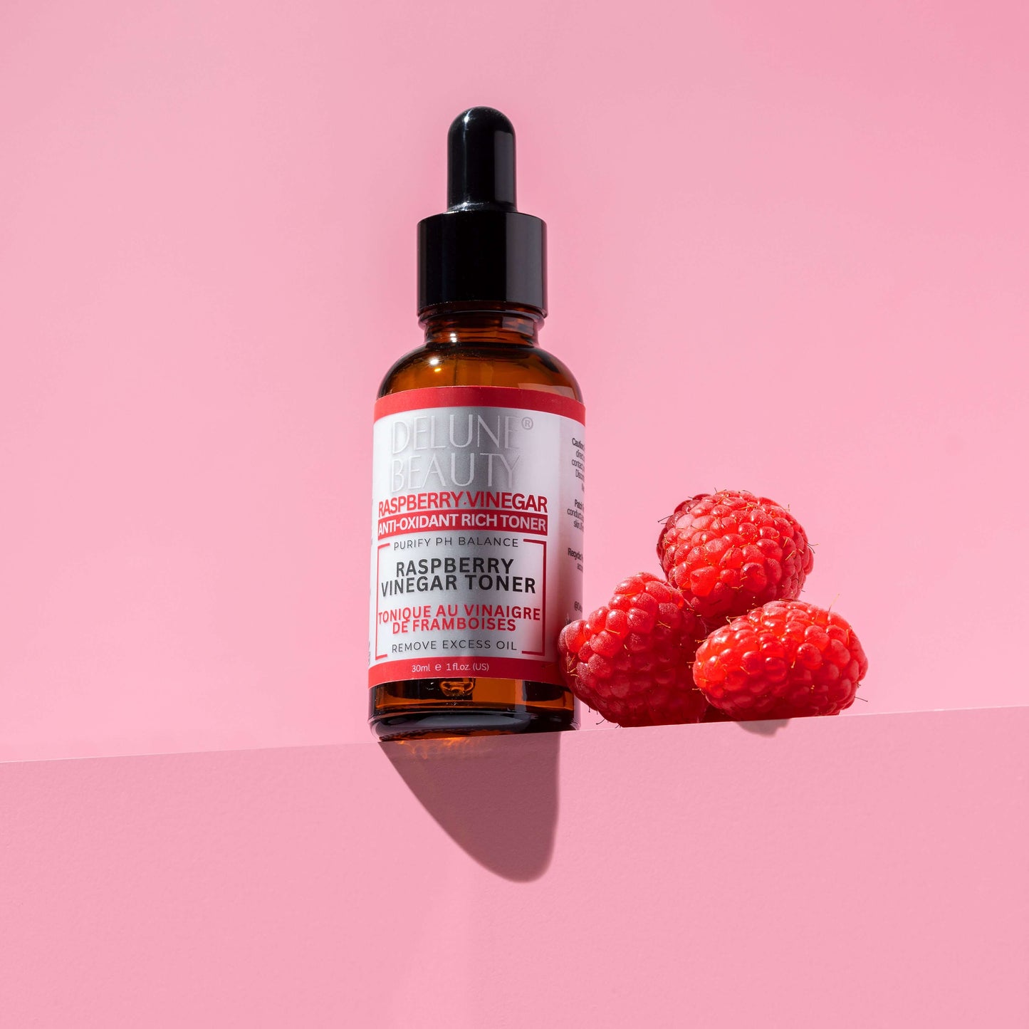 Delune Raspberry Vinegar Toner bottle next to fresh raspberries on a pink background; skincare for oily and blemish-prone skin.