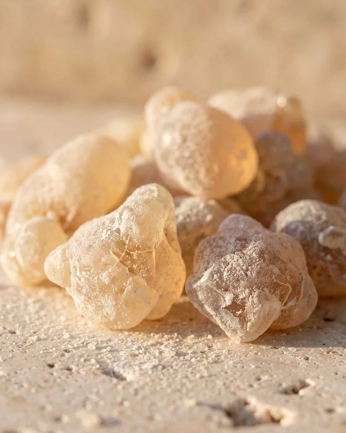Raw frankincense resin tears glowing honey-amber in warm backlight on bone travertine — the warm, resinous scent of Delune Frankincense Pure Essential Oil