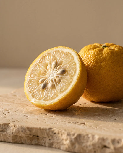 A halved yuzu beside a whole bumpy-skinned yuzu on warm travertine — the bright, floral citrus scent of Delune Yuzu Pure Essential Oil